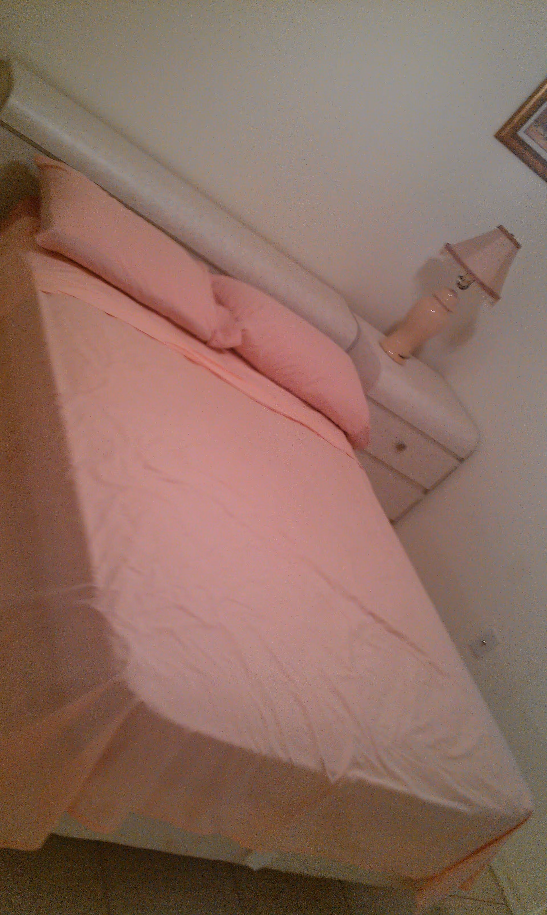 A bedroom with a bed made up with pink sheets and pillows. There is a white padded headboard and a white nightstand with a pink lamp on it. A framed picture is partially visible on the wall above the nightstand.