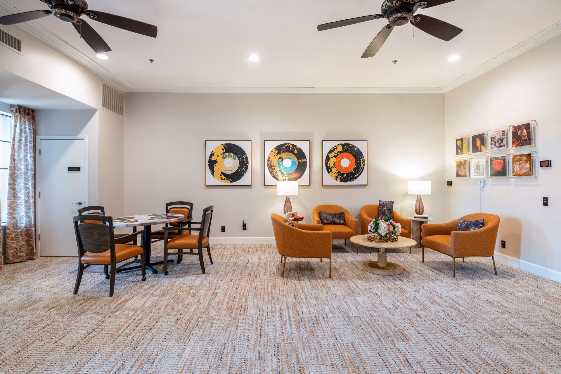 A spacious and well-lit common area with a seating arrangement consisting of four orange armchairs around a round wooden coffee table with a floral centerpiece. To the left, there is a small round table with four chairs. The walls are decorated with three framed abstract art pieces and a display of vinyl record covers. The room has ceiling fans and recessed lighting, with patterned curtains on the left side.