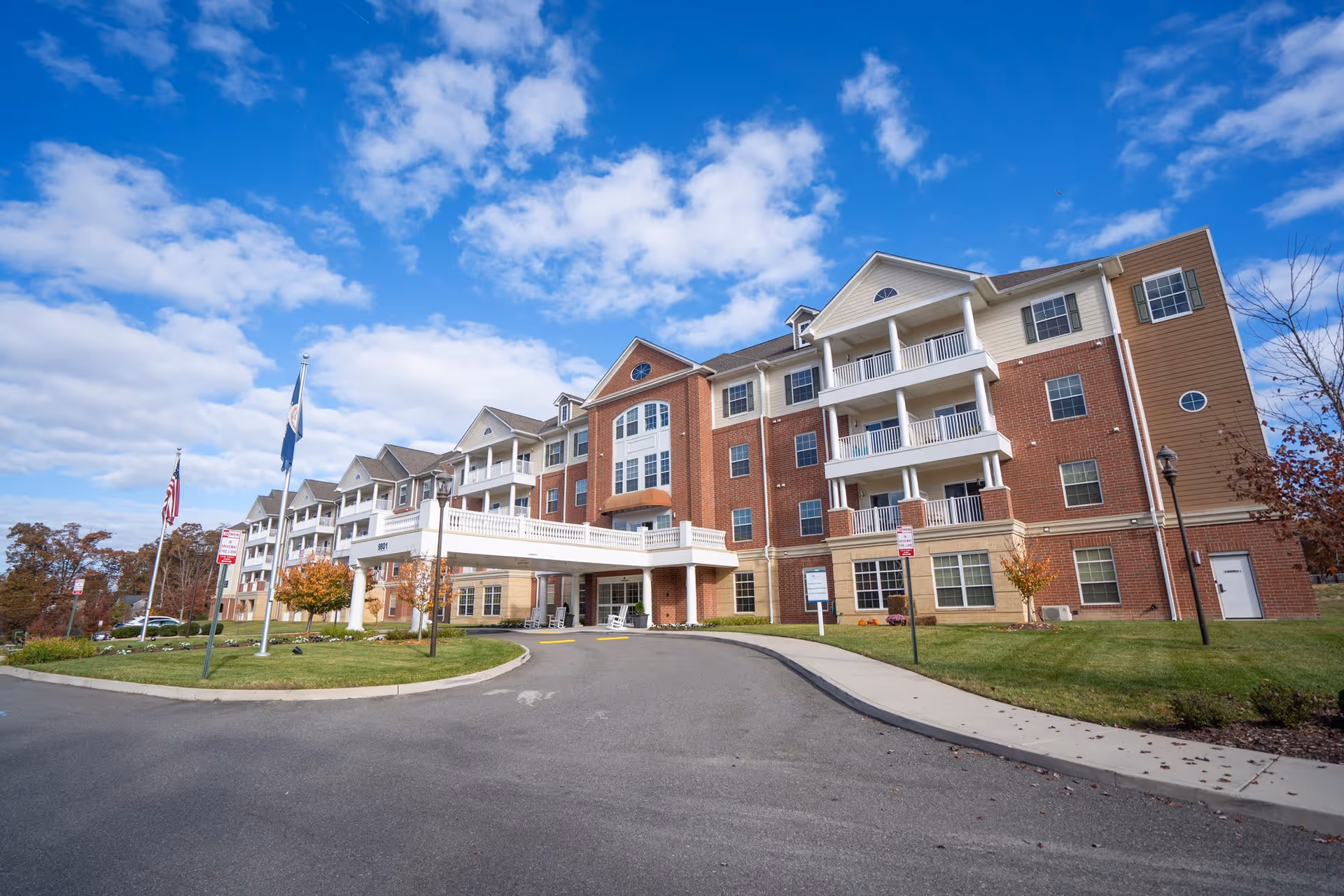 Exterior view of a multi-story senior living facility building with a covered entrance, balconies, and a driveway. The building features a combination of red brick and beige siding under a partly cloudy blue sky. There are flags and small trees planted in the landscaped area near the entrance.