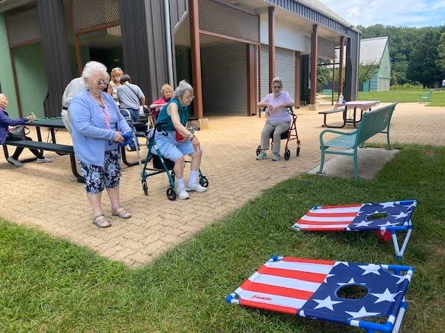 A group of elderly women, some using walkers, are gathered outside near a building with picnic tables and benches. Two cornhole boards decorated with an American flag design are set up on the grass in the foreground. The setting appears to be a sunny day with green grass and trees in the background.