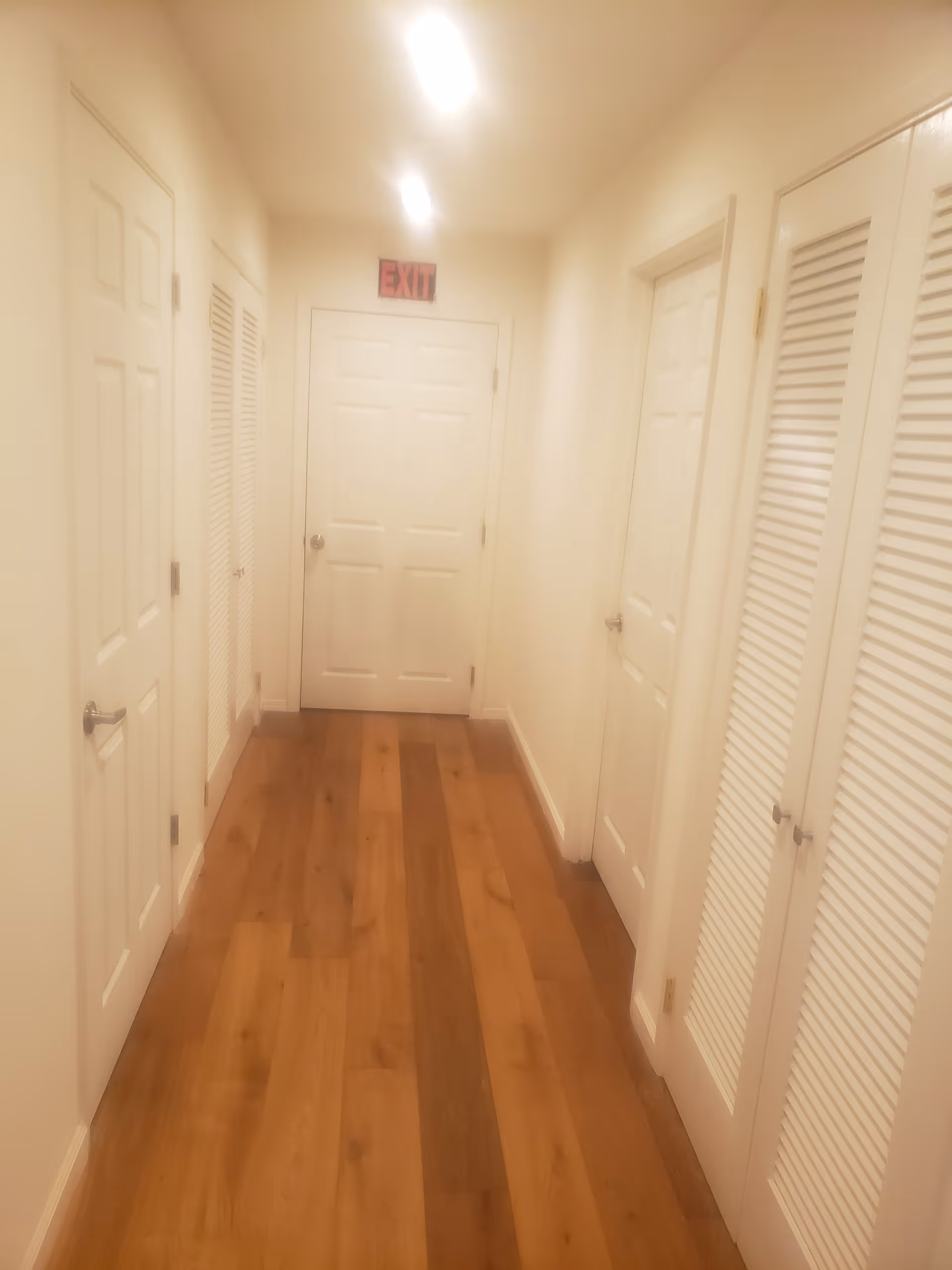 A hallway with wooden flooring and white walls, featuring multiple white doors on both sides and an exit sign above a door at the end of the hallway.