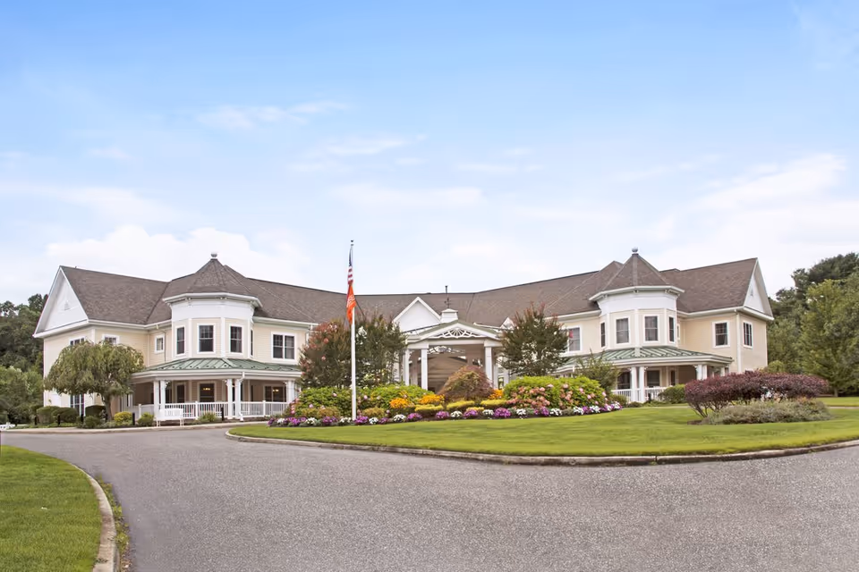 Front exterior of a two-story senior living building with a circular driveway, flagpole, and landscaped flower beds.