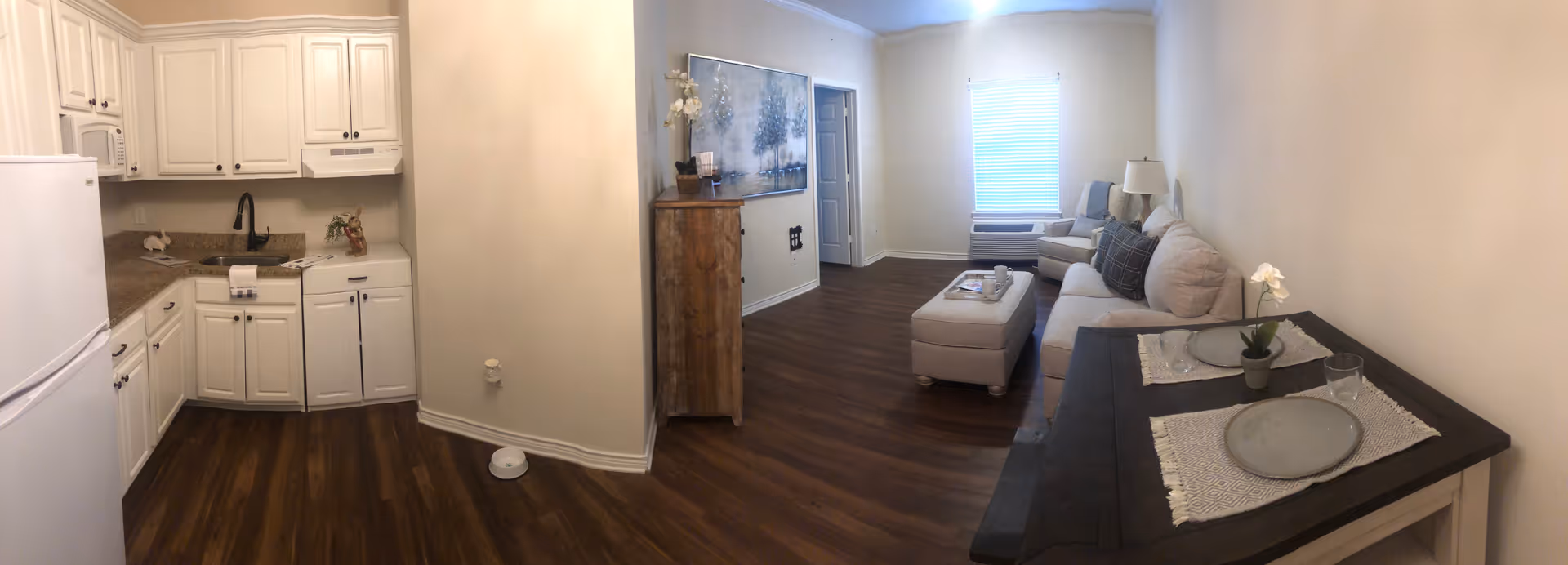 Open-plan interior showing a small white kitchen on the left and a living area with a sofa, ottoman and table on the right.