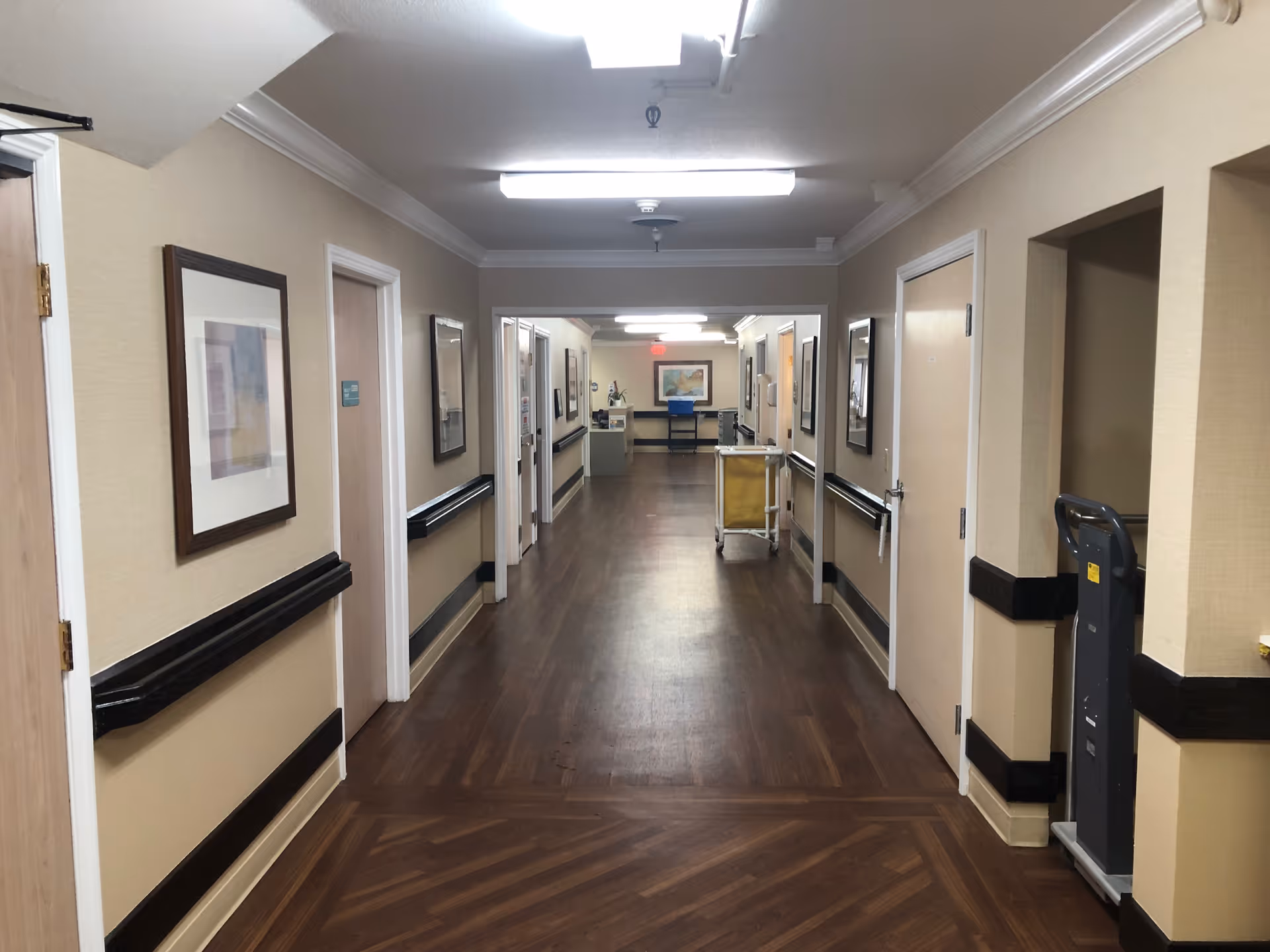 A long, well-lit hallway in a healthcare facility with wooden flooring and beige walls. Several doors line both sides of the corridor, with framed artwork hanging on the walls. Handrails run along the walls on both sides. At the far end, there is a cart and a framed picture on the wall.