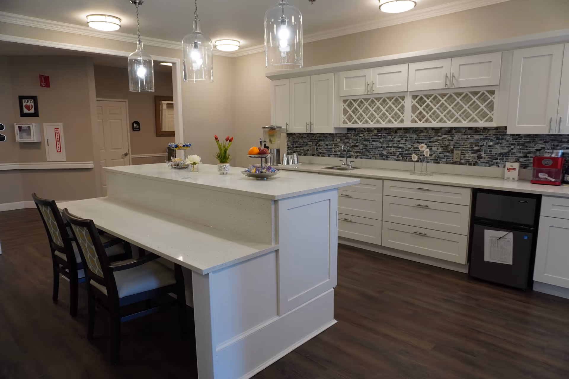 A modern kitchen area in a senior living facility with white cabinetry, a mosaic tile backsplash, and a large island with a raised counter and seating for two. The island has decorative items including flowers and a fruit bowl. Pendant lights hang above the island, and there is a small refrigerator and coffee machine on the right side.