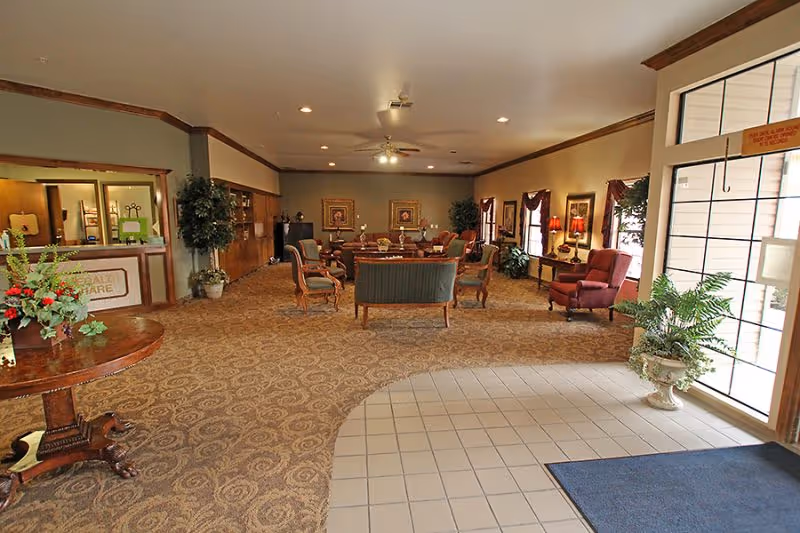 Spacious senior living facility lobby with a reception desk, multiple seating areas, plants, and large windows at the entrance.