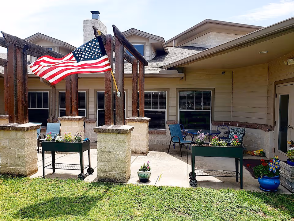 Outdoor patio area of an assisted living facility with an American flag mounted on wooden beams, two raised garden beds with flowers, patio chairs, and a small table. The building exterior features beige siding and multiple windows.