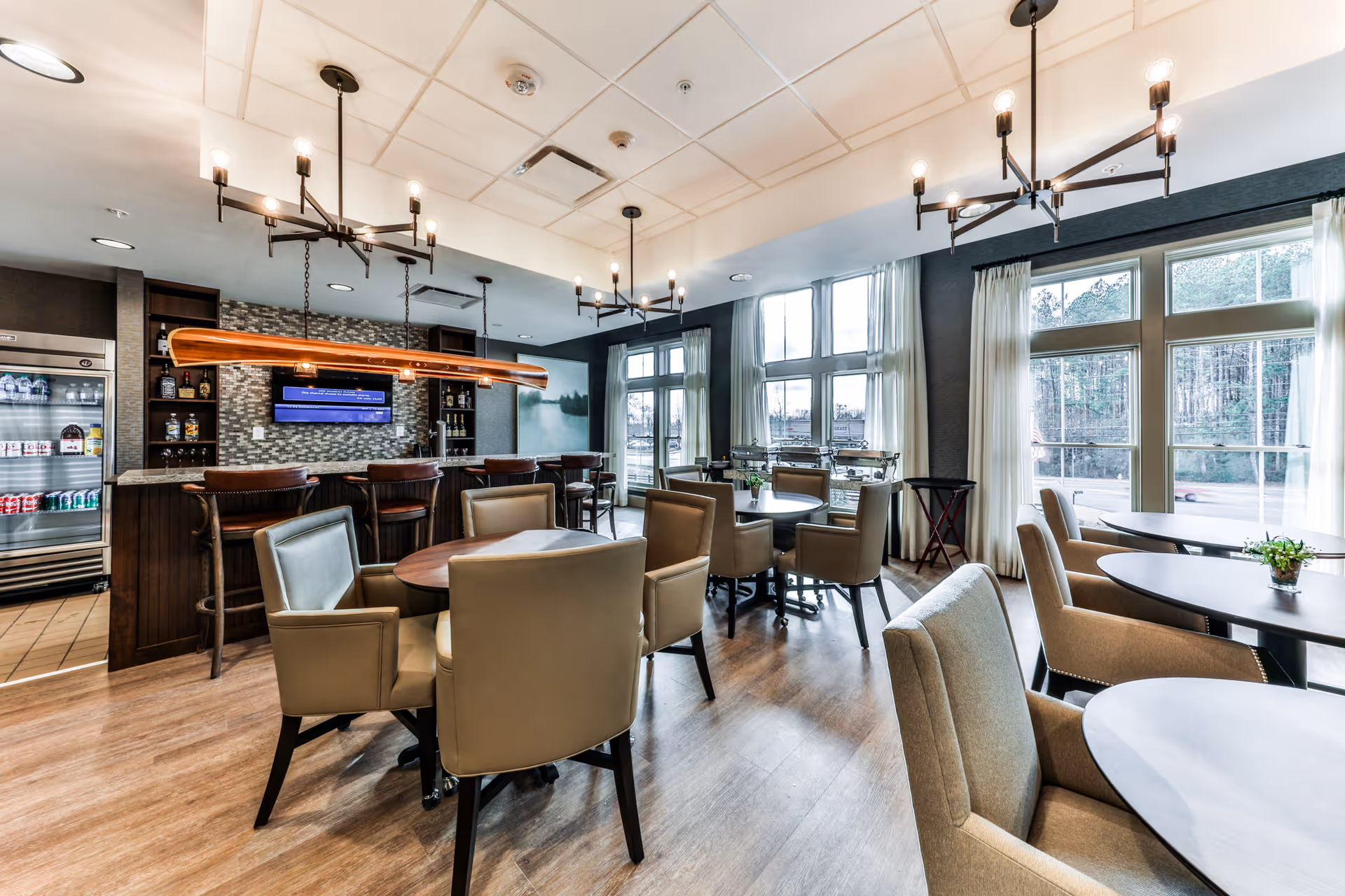 A bright and modern dining area with multiple round tables surrounded by beige upholstered chairs. Large windows with white curtains allow natural light to fill the room. There is a bar area with high chairs, a refrigerator stocked with beverages, and a wall-mounted TV above the bar. Contemporary chandeliers hang from the ceiling, and the floor is wood.