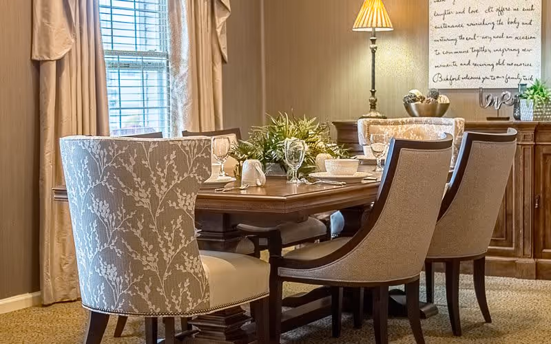 A dining room with a wooden table set for a meal, featuring six upholstered chairs with patterned and plain fabric. The table is decorated with glassware, white dishes, and a green centerpiece. In the background, there is a window with beige curtains, a lamp on a sideboard, and a framed wall decoration with text.