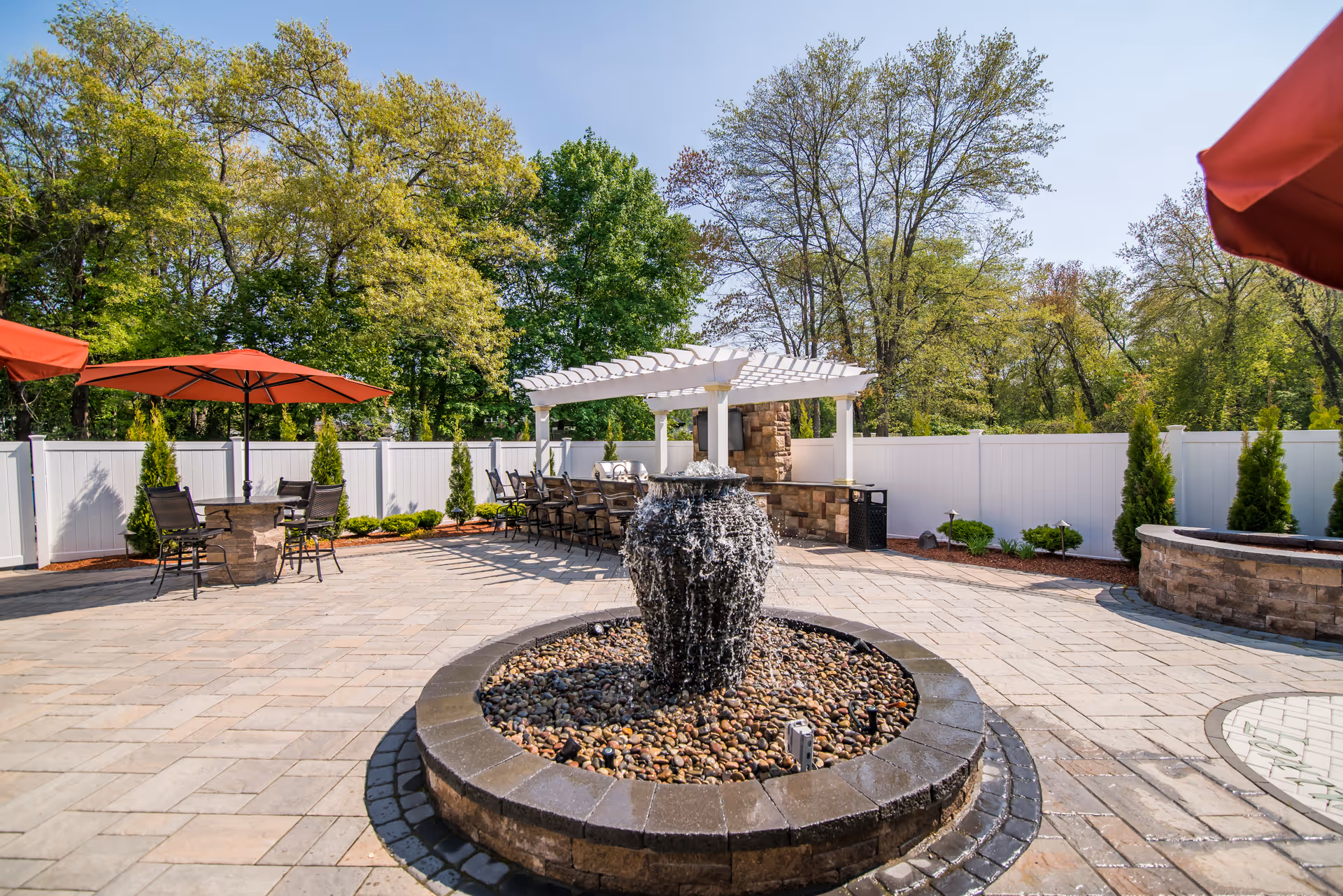 Outdoor patio area with a central stone water fountain surrounded by a circular stone border filled with pebbles. There are several seating areas with tables and chairs, some shaded by red umbrellas. A white pergola with additional seating and a stone fireplace is visible in the background, enclosed by a white fence and surrounded by trees.