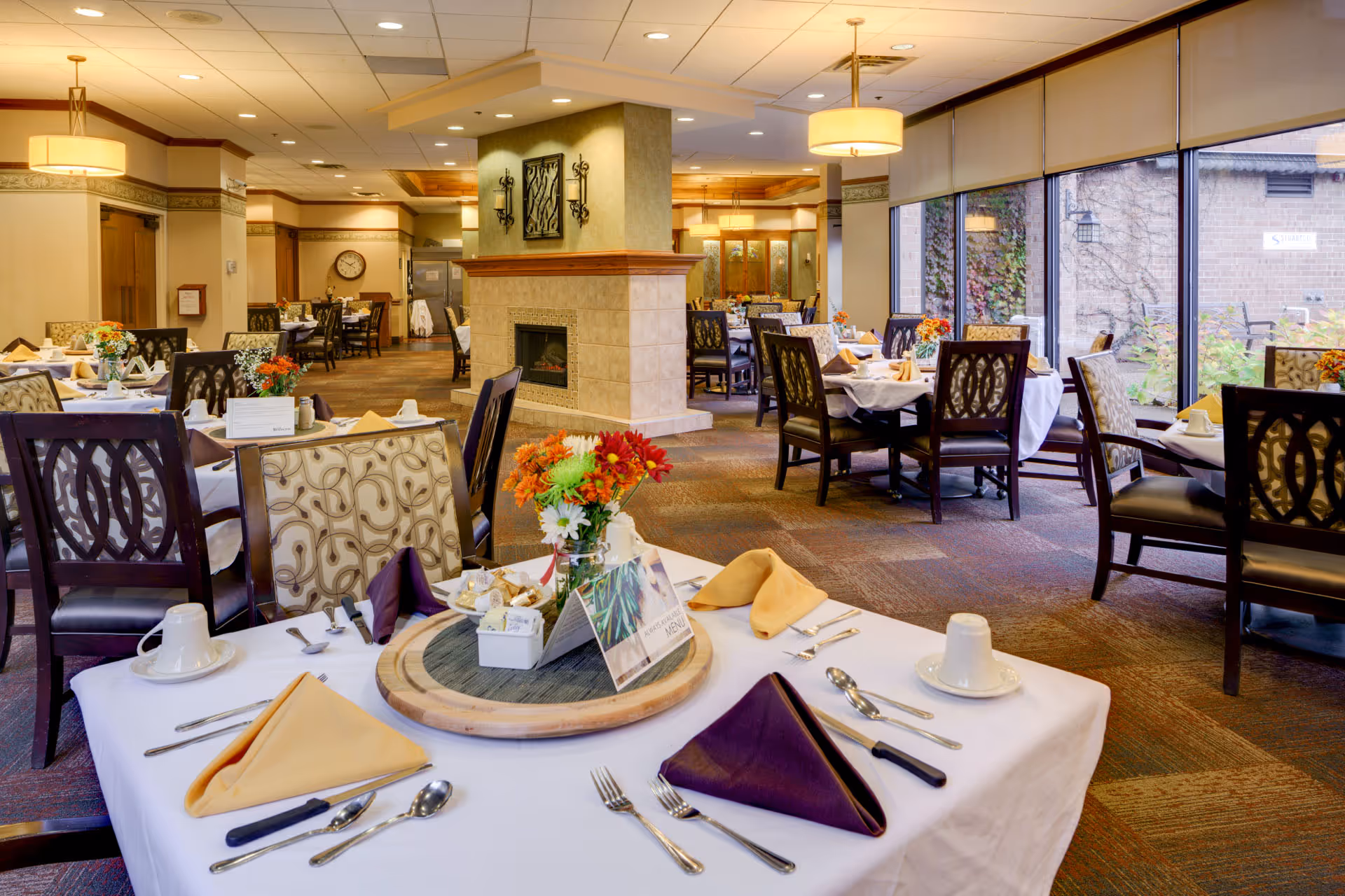 Bright, well-appointed dining room with multiple set tables, a central fireplace, large windows, and floral centerpieces.