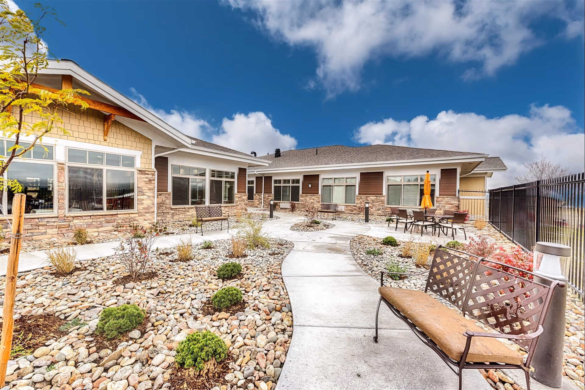 Outdoor courtyard area of a senior living facility with a paved walkway, benches, small shrubs, and patio tables with chairs and an orange umbrella. The building has large windows and stone and wood siding under a partly cloudy blue sky.