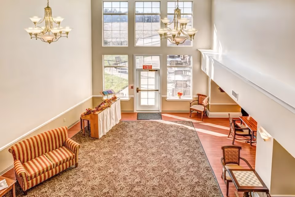 Bright and spacious interior common area with large windows letting in natural light. The room features a patterned carpet, a striped sofa, a small table with a chessboard, a wooden desk with chairs, and two chandeliers hanging from the ceiling. There is an exit door with windows above and on the sides, and a small table with decorative items near the door.