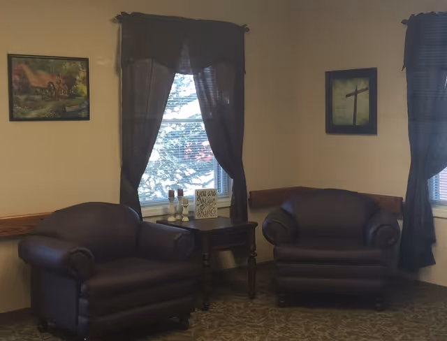 Two dark armchairs flank a small wooden side table beneath curtained windows in a cozy sitting area.