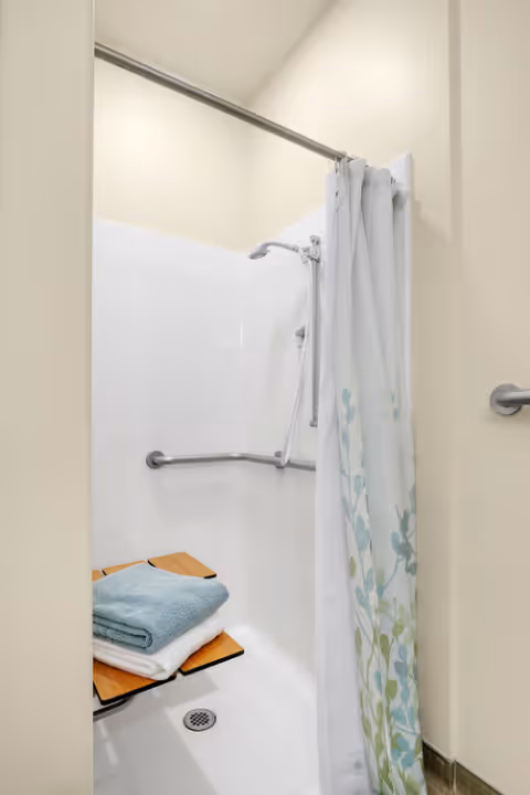 A shower area with a white wall surround, a metal grab bar, and a foldable wooden shower seat holding two folded towels, one blue and one white. A shower curtain with a floral pattern is partially drawn to the side.