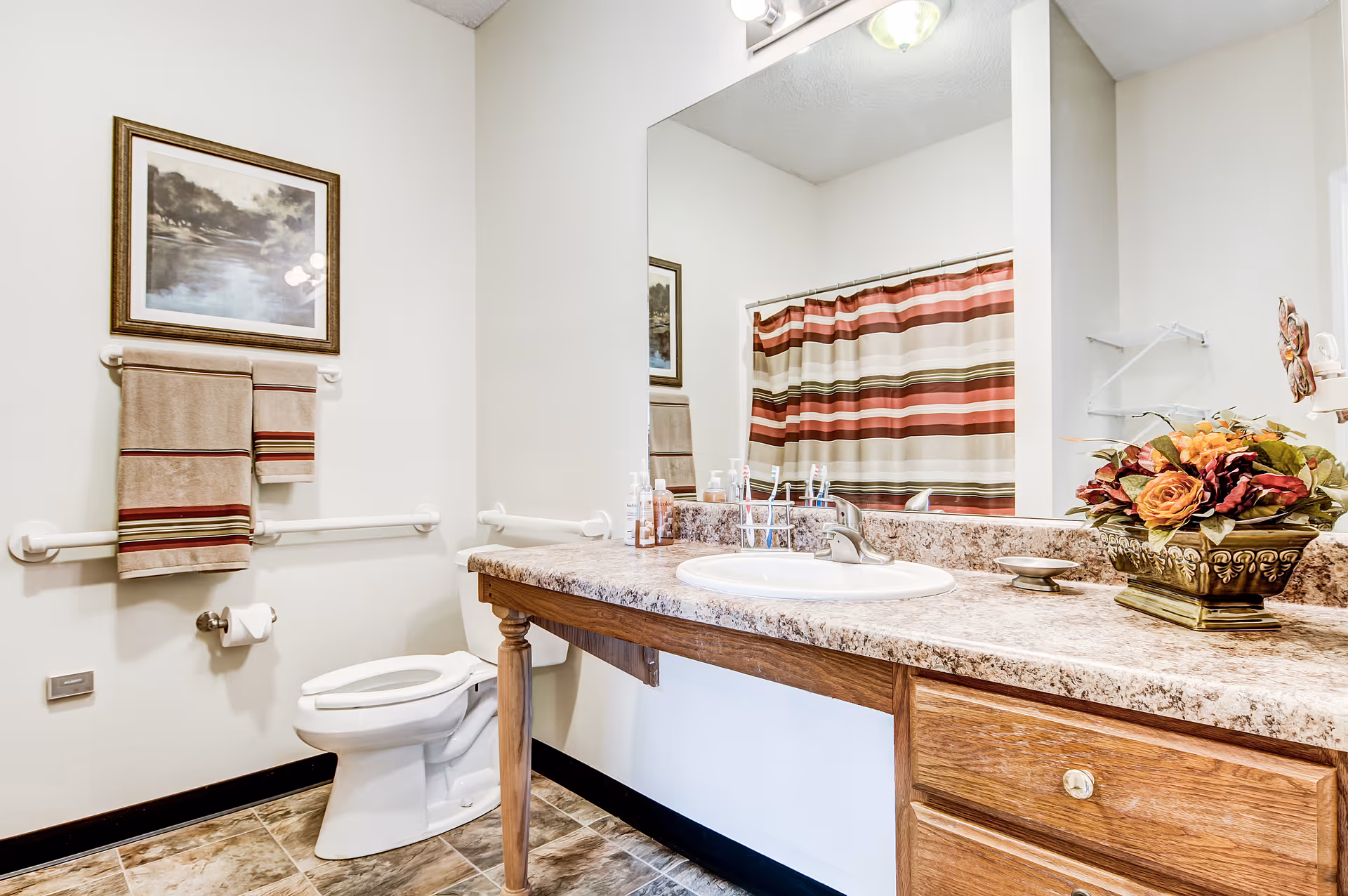 A clean bathroom with a toilet, a large mirror above a countertop with a sink, and a striped shower curtain. The countertop has a decorative flower arrangement and several toiletries. There are two towels hanging on a wall-mounted towel rack beneath a framed landscape painting. The floor has tile flooring and the walls are painted white.