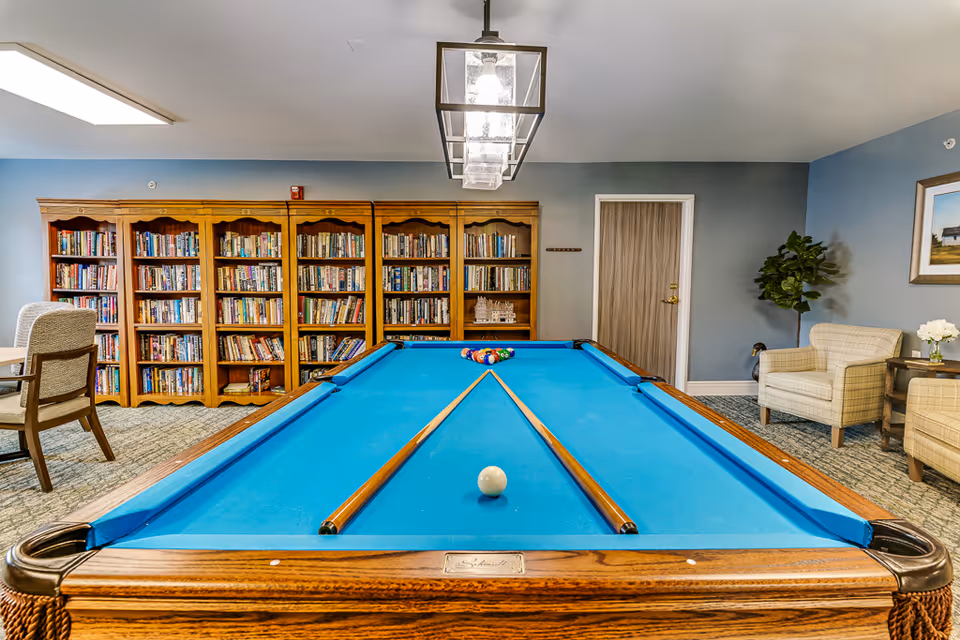 A recreational room featuring a pool table with two cues resting on the blue felt surface. Behind the pool table is a large wooden bookshelf filled with books. To the right, there are two beige armchairs, a small side table with a vase of white flowers, a potted plant, and a framed picture on the blue wall. A modern rectangular light fixture hangs from the ceiling above the pool table.
