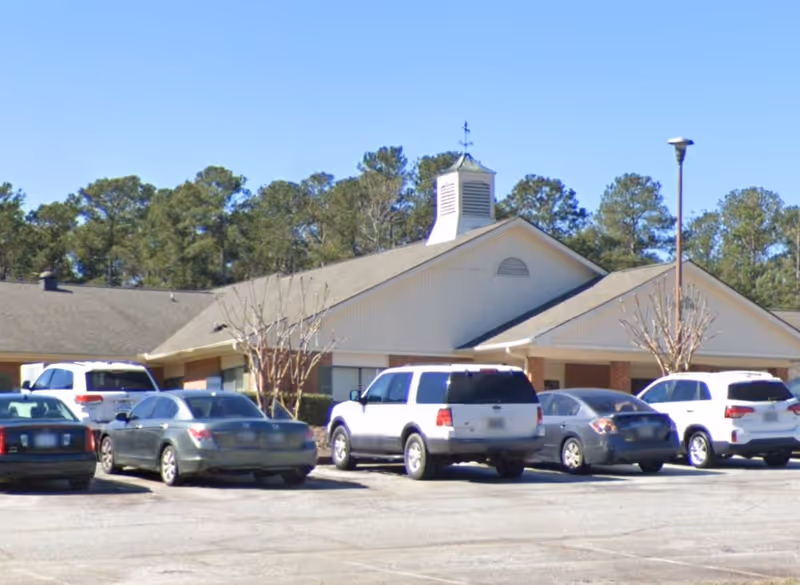 Exterior view of McCormick Post-Acute facility showing a single-story building with a pitched roof, a cupola on top, several parked cars in front, and trees in the background under a clear blue sky.
