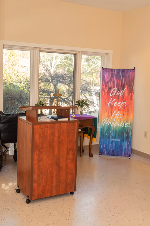 A small chapel or prayer room with a wooden podium on wheels, a table with flowers and candles, and a colorful banner that reads 'God Keeps His Promises' with a Bible reference to Deuteronomy 7:9. Large windows show trees outside.