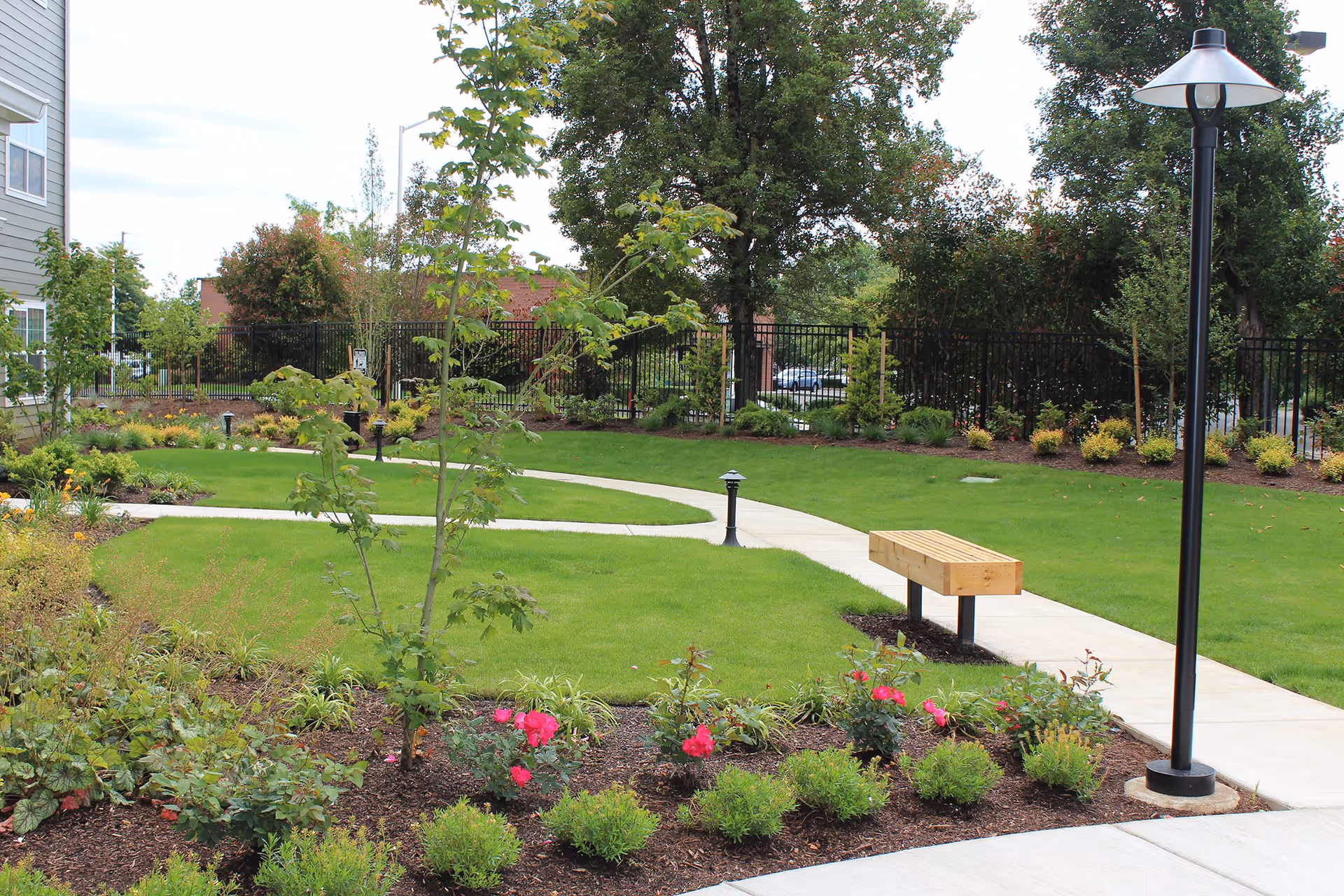 A landscaped outdoor garden area with a curved concrete walkway, green grass, various plants and flowers, a wooden bench, and a tall black lamp post. Trees and shrubs surround the area, and a black metal fence is visible in the background.