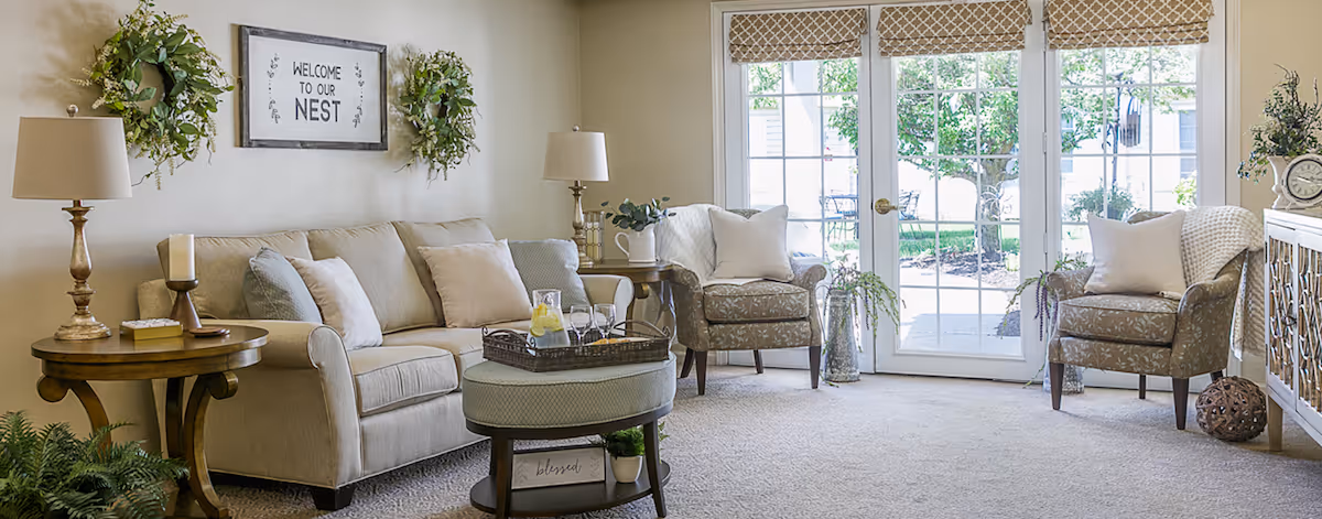 Bright, cozy living room with a sofa, armchairs, coffee table, lamps, decorative wreaths, and French doors opening to a garden.