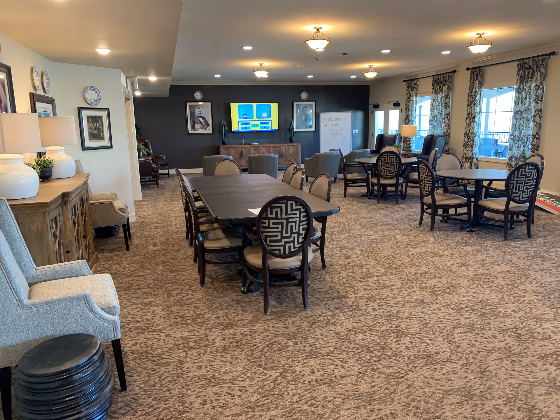 A spacious common area with multiple tables and chairs arranged for group seating. The room has carpeted flooring, several large windows with patterned curtains allowing natural light, and a dark accent wall with a mounted TV and framed pictures. There are also lamps and decorative items on a wooden sideboard along one wall.
