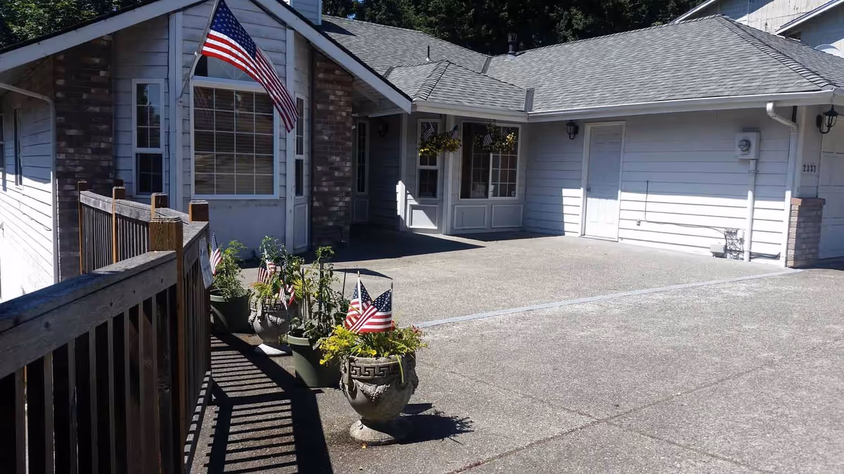 Exterior view of a single-story senior care facility building with a concrete driveway and a wooden ramp. Several potted plants with small American flags are placed along the ramp. The building has white siding, a brick accent wall, and an American flag mounted near a large window.