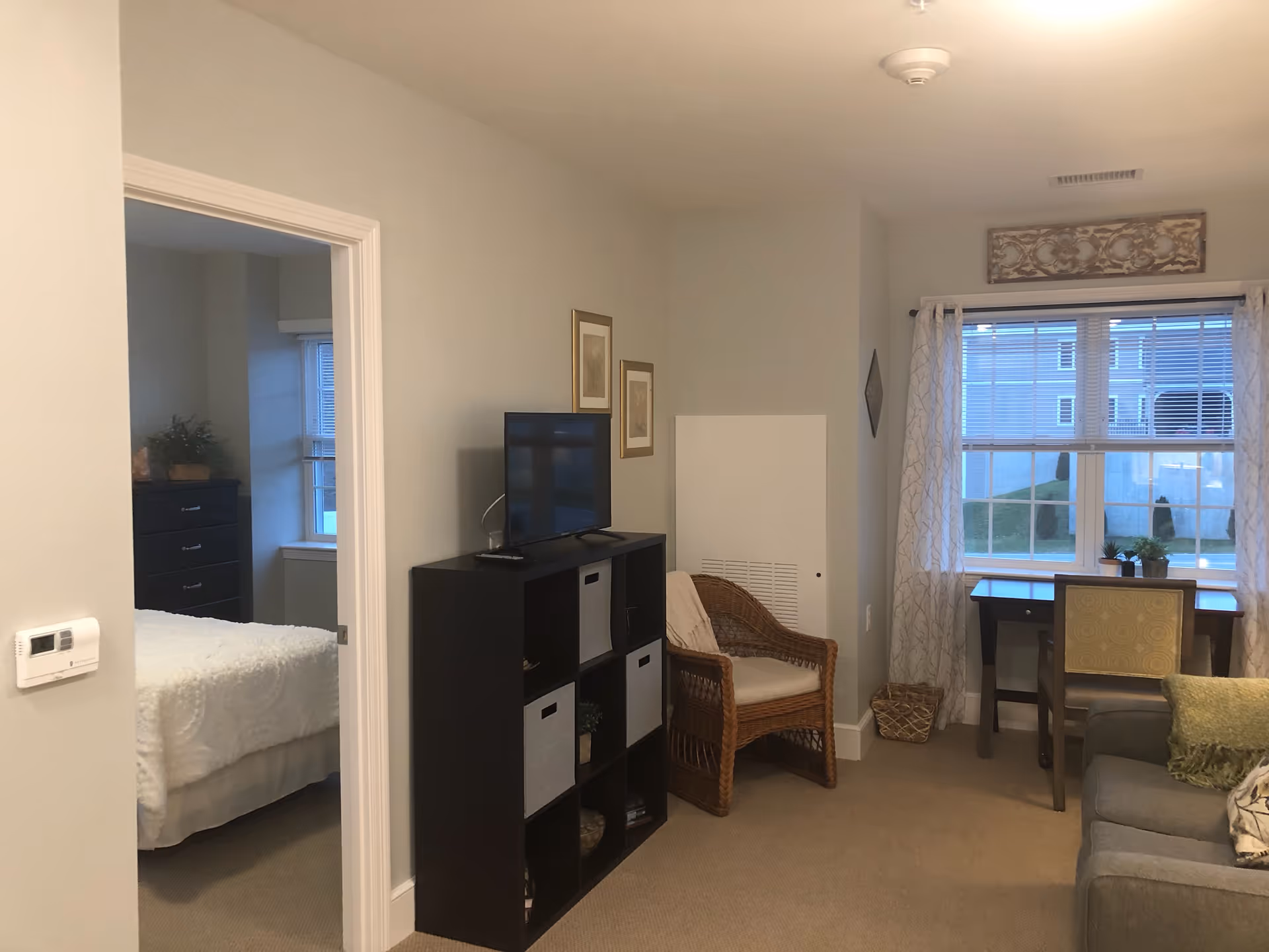 Cozy living room showing a TV on a shelving unit, a wicker chair, a desk by a window, and a view into an adjacent bedroom.
