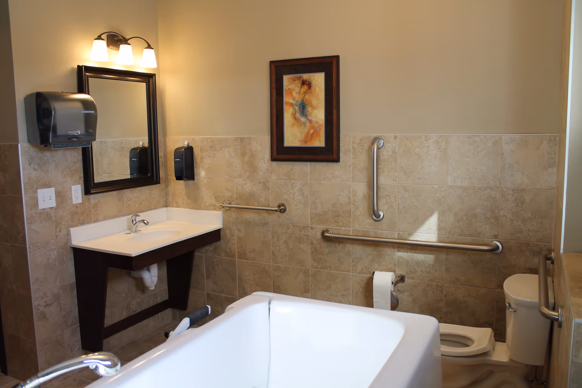 A bathroom with beige tiled walls and floor, featuring a white bathtub in the foreground, a white sink with a dark wooden base and a mirror above it on the left wall. There are two black soap or paper towel dispensers mounted on the wall near the sink. The toilet is visible on the right side with grab bars installed on the walls for accessibility. A framed abstract painting hangs on the wall above the grab bars. The lighting fixture above the mirror has three lights.