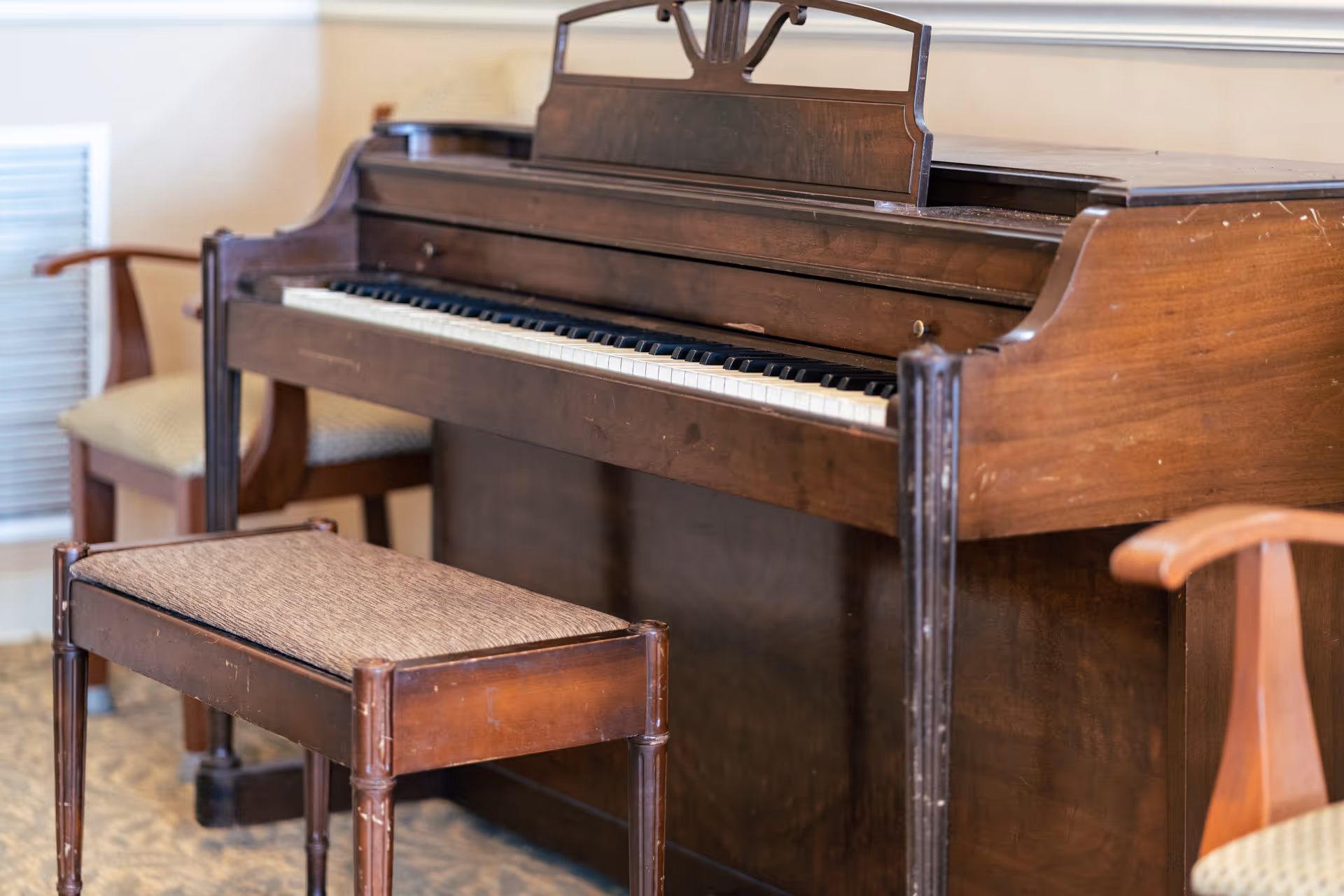A wooden upright piano with a matching cushioned bench in front of it, surrounded by wooden armchairs with beige cushions in a softly lit room.