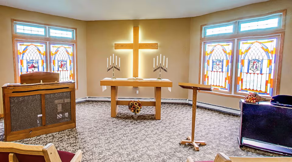 Interior of a small chapel room with a wooden cross on the wall illuminated from behind. There are two stained glass windows on each side of the cross, a wooden altar table with two candelabras and an open book, a wooden lectern, and a piano. The floor is carpeted with a floral pattern.