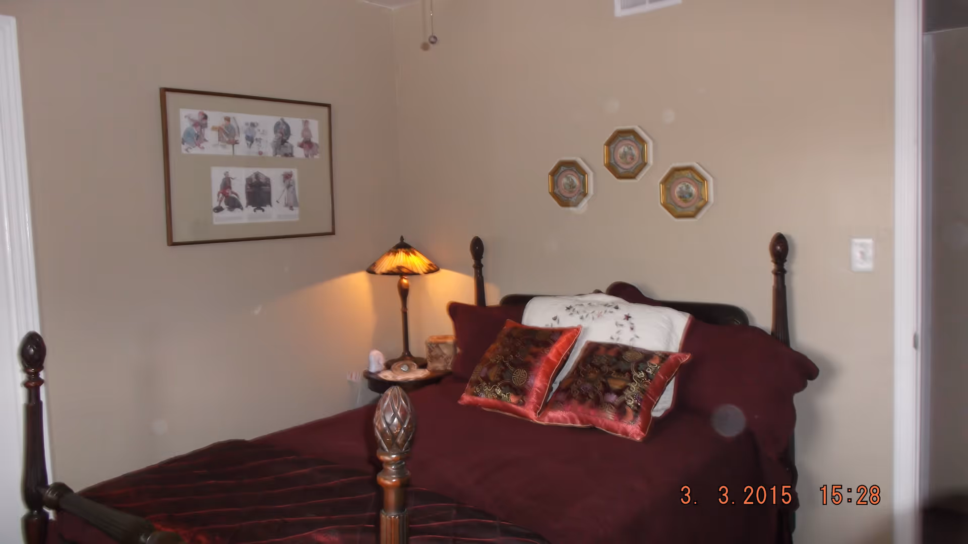 A bedroom with a dark wooden four-poster bed covered with a burgundy bedspread and decorative pillows. A small bedside table with a lamp and a framed picture on the wall are visible. The walls are painted beige.