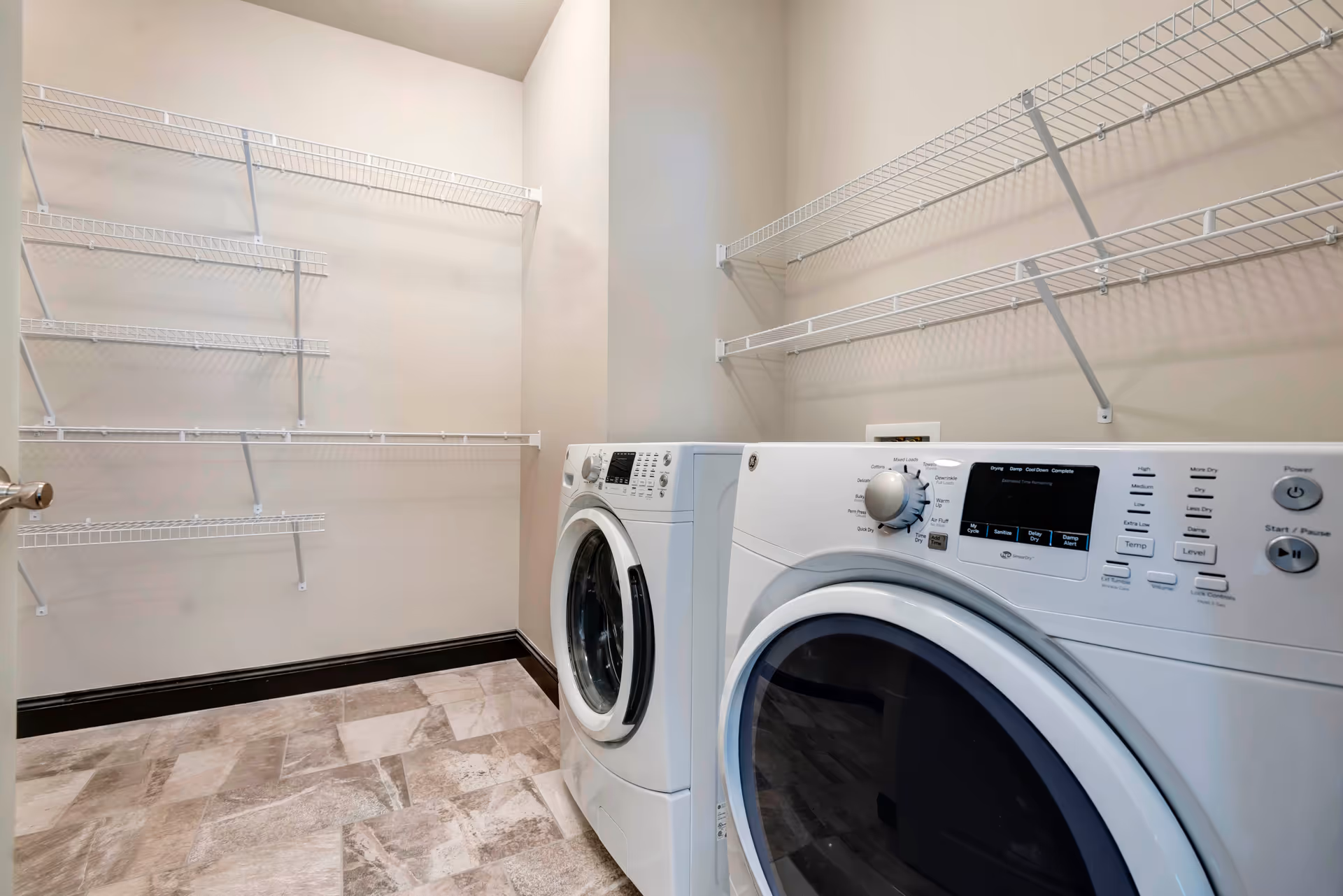 Laundry room with a front-loading washing machine and dryer side by side, beige walls, multiple white wire shelves mounted on the walls, and a tiled floor.