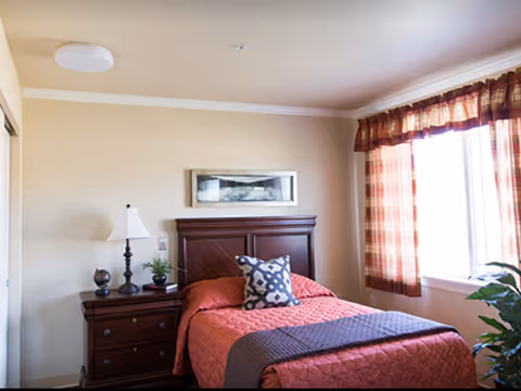 A cozy bedroom with a single bed covered in a red quilt and a decorative pillow. Next to the bed is a wooden nightstand with a lamp, a small plant, and a decorative item. A framed picture hangs above the headboard. The room has a window with plaid curtains allowing natural light to enter.