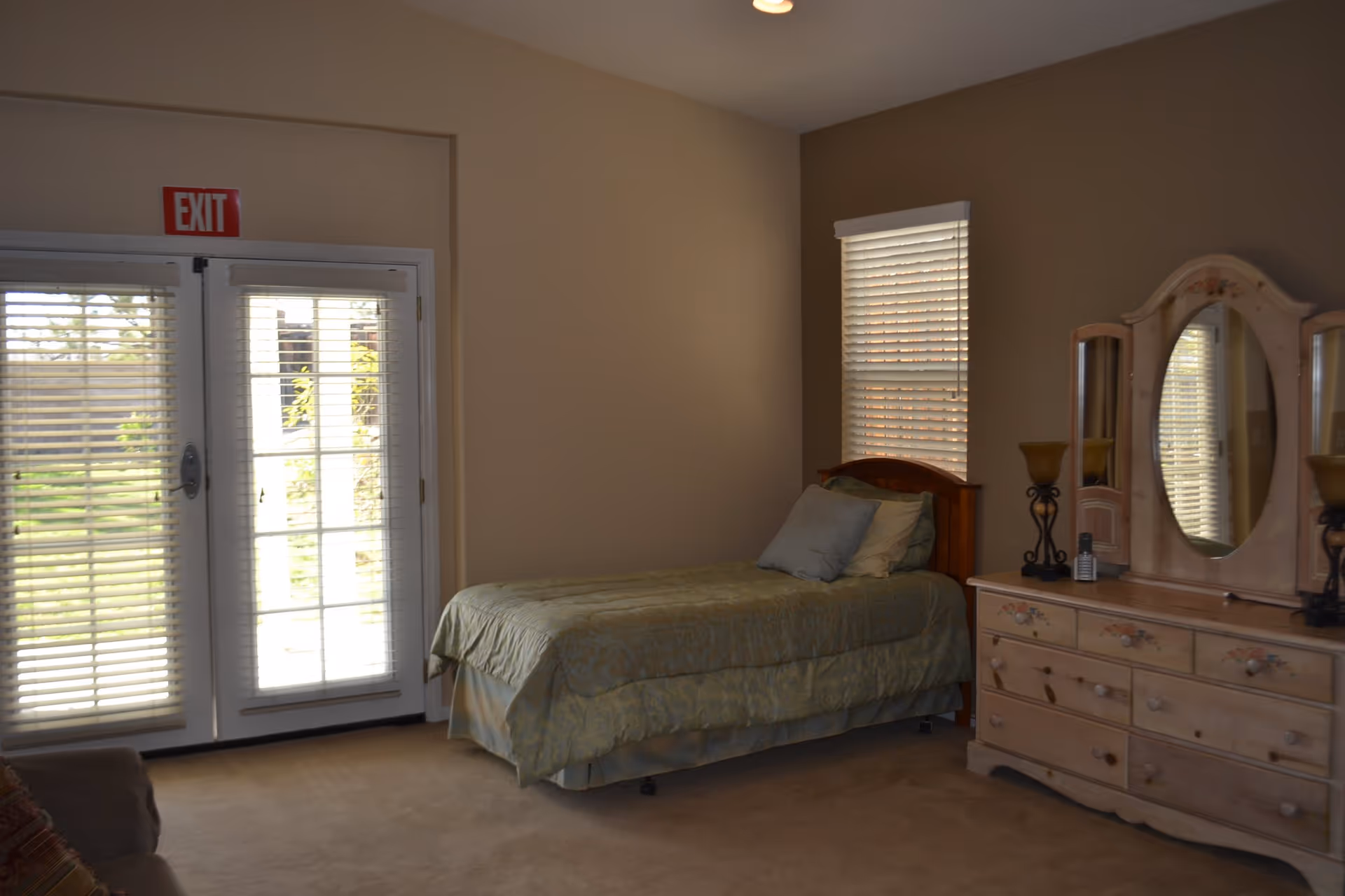 A bedroom with a single bed, wooden dresser and mirror, window blinds, and glass French doors with an EXIT sign above.