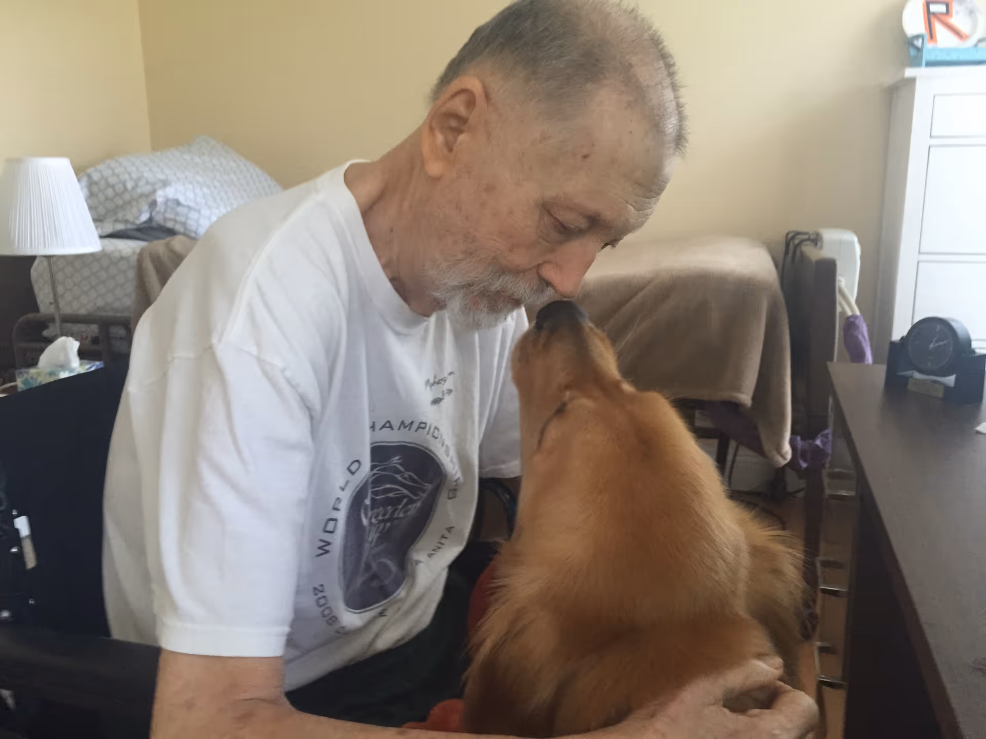 An elderly man sitting in a wheelchair inside a room, affectionately interacting with a golden retriever dog that is close to his face.