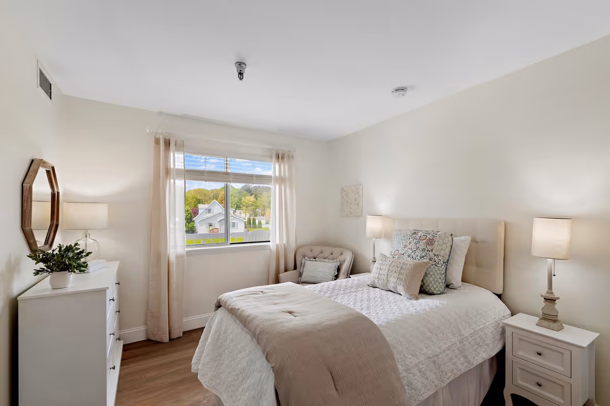 A bright and cozy bedroom with a neatly made bed featuring multiple pillows and a beige throw blanket. There is a white dresser with a plant and a mirror on the left side, a window with sheer curtains letting in natural light, a cushioned armchair in the corner, and two bedside tables each with a lamp. The walls are painted light beige and the floor is wooden.