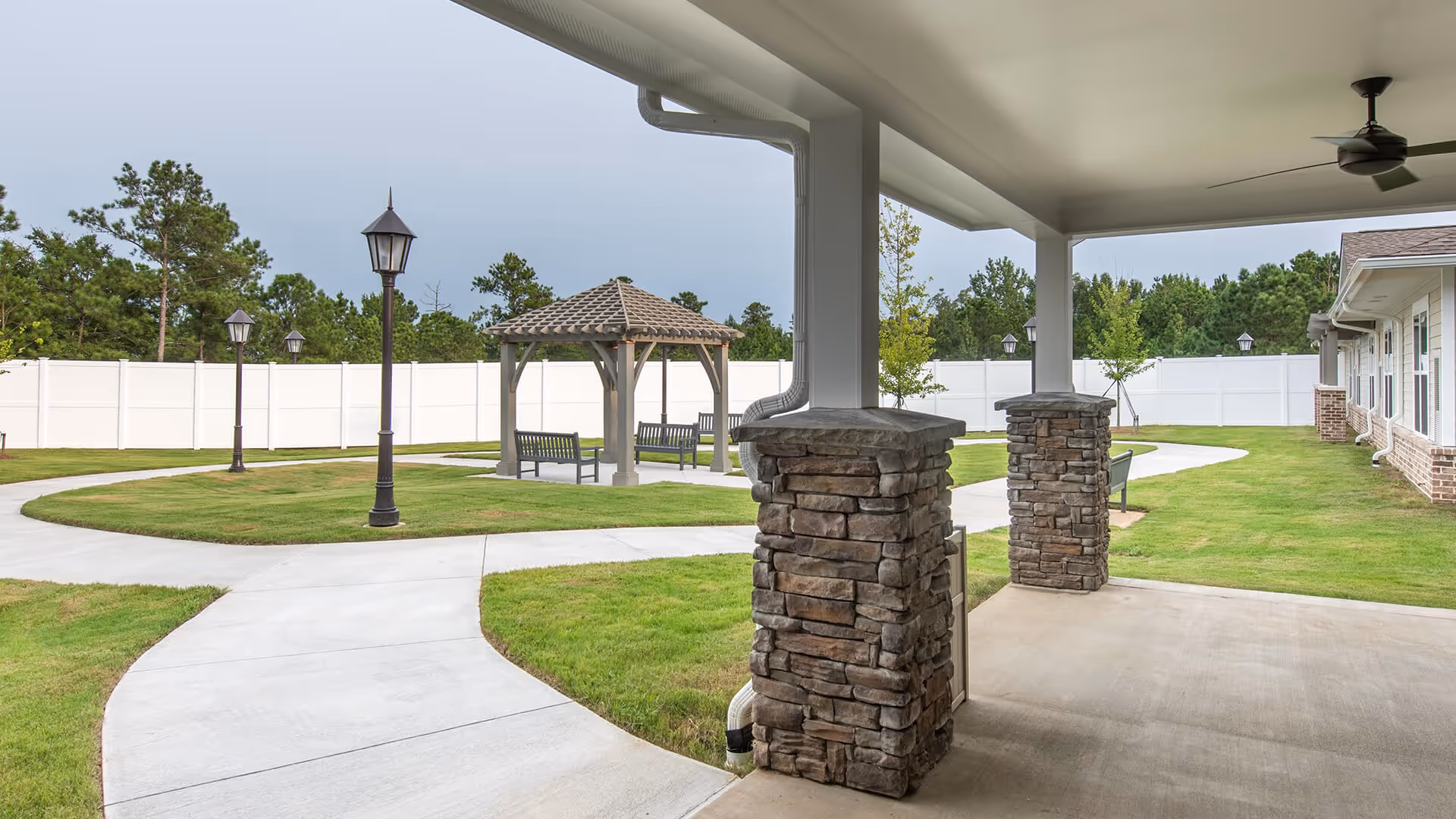 Covered patio with stone columns overlooking a paved walkway, gazebo, benches, lamp posts and a fenced grassy courtyard.