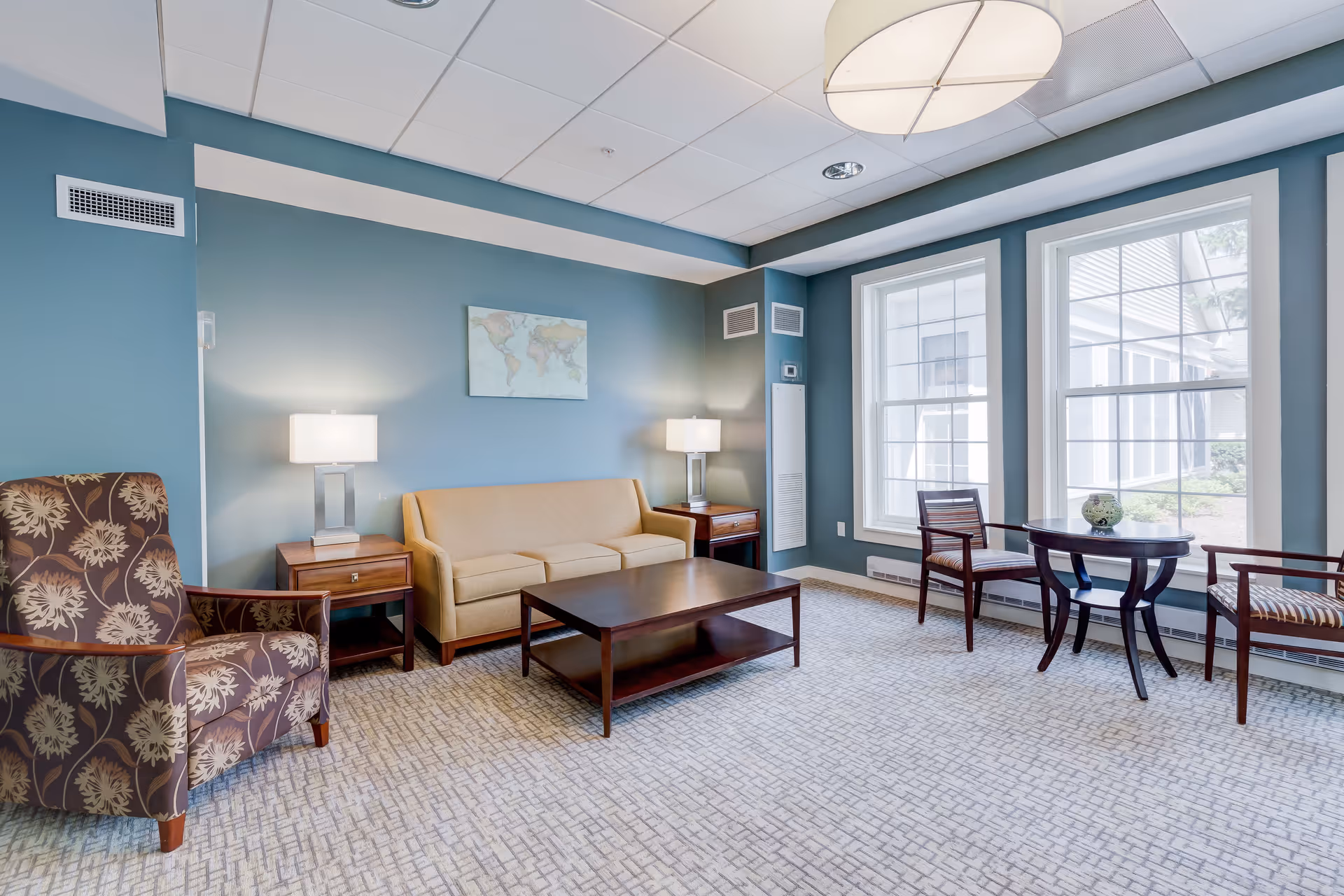 A bright and cozy living room with teal walls, a beige sofa flanked by two wooden side tables with lamps, a patterned armchair, a wooden coffee table, and a small round table with two chairs near large windows letting in natural light.