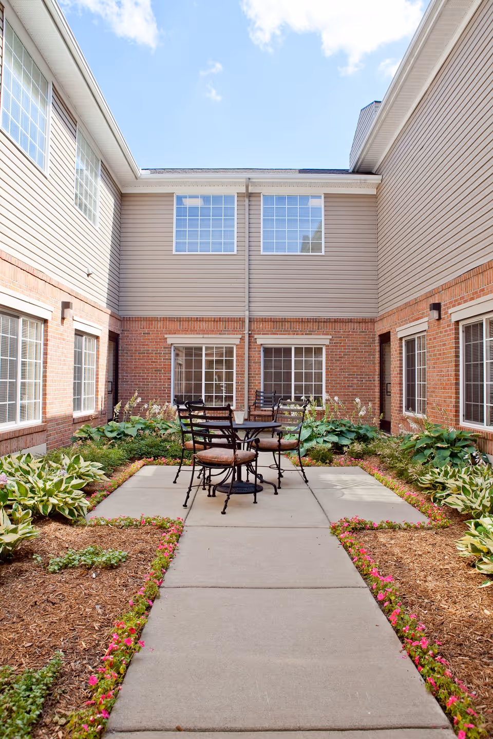 Outdoor courtyard area with a concrete pathway leading to a round metal table surrounded by four cushioned chairs. The courtyard is bordered by flower beds with green plants and small pink flowers. The surrounding building has beige siding on the upper half and red brick on the lower half with multiple windows.