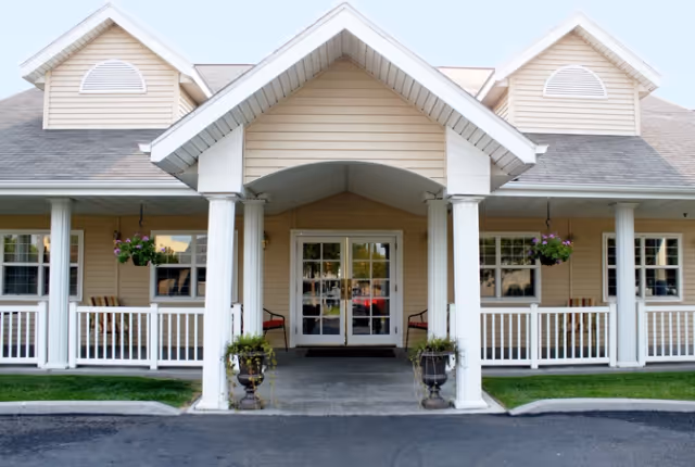 Front exterior view of a senior living facility with beige siding, white columns, and a covered entrance. There are hanging flower baskets and potted plants near the entrance, with a paved driveway in front.