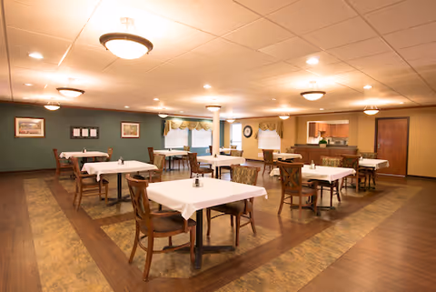 Spacious dining room with several tables covered in white tablecloths, wooden chairs, warm overhead lighting, and a serving counter.