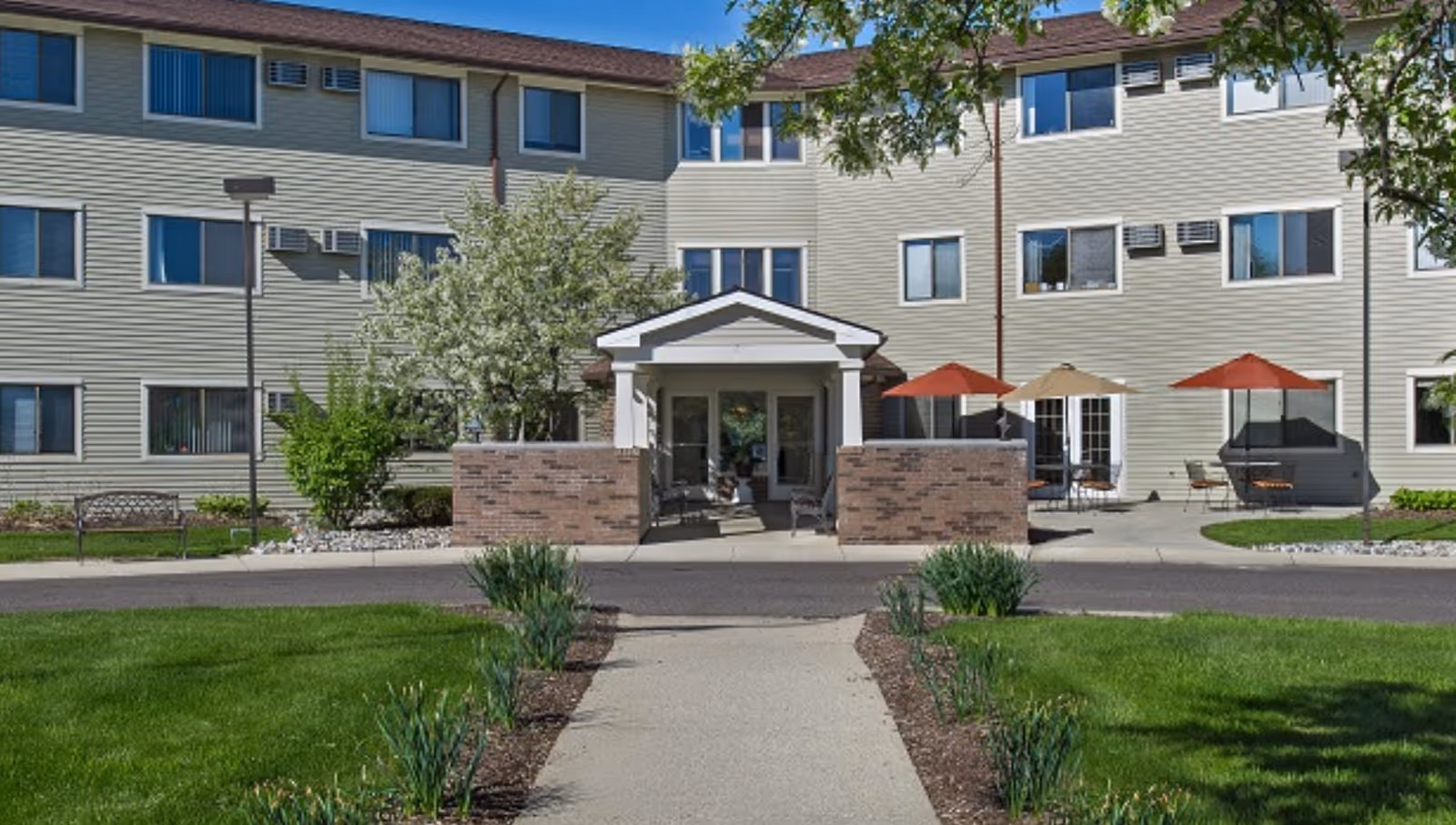 Two-story beige senior living building with glass entrance and patio seating under umbrellas.