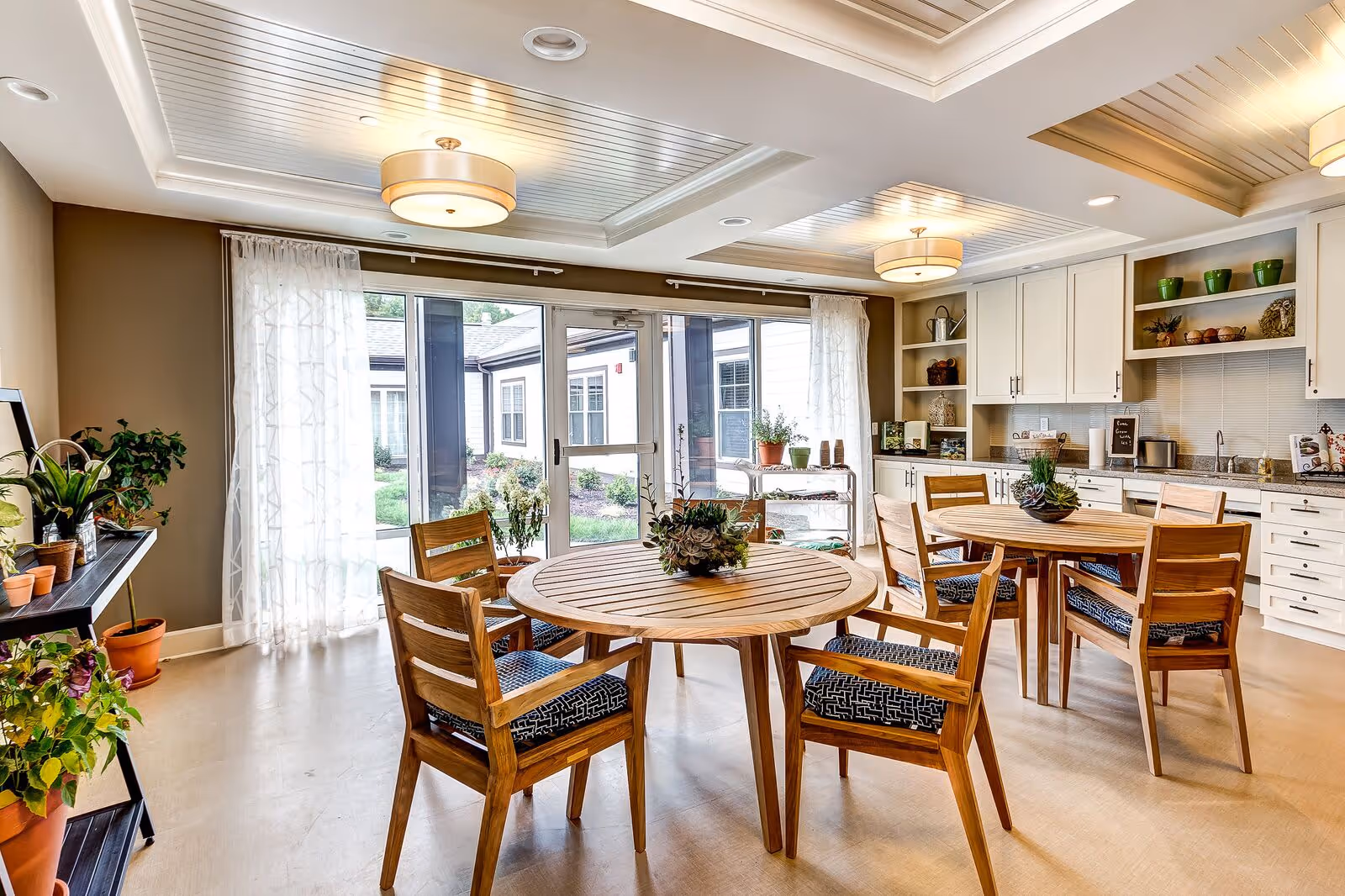 A bright and airy dining area with two round wooden tables, each surrounded by four wooden chairs with patterned cushions. The room features large glass doors leading outside, sheer white curtains, and a ceiling with recessed lighting and two modern light fixtures. There are white cabinets and shelves along one wall with decorative items and plants, and additional potted plants on a black metal shelf and on the floor.
