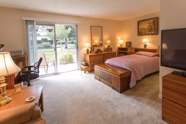 A well-lit bedroom with a large bed covered in a pink quilt, wooden dressers with lamps and framed pictures, a flat-screen TV on a wooden stand, a sliding glass door leading to a garden patio, and a comfortable chair with a side table and lamp.