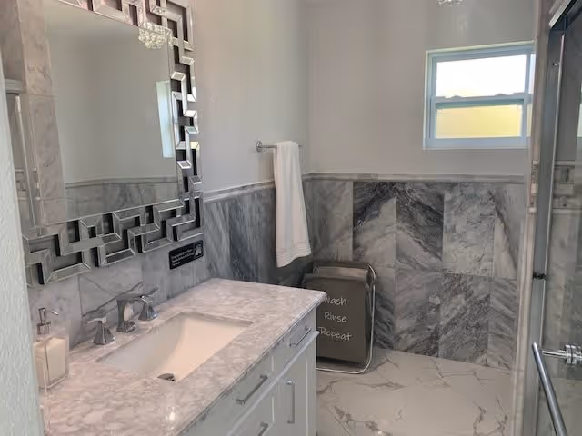 A modern bathroom with marble walls and floor tiles. There is a white vanity with a marble countertop and an under-mount sink. Above the sink is a decorative mirror with a geometric frame. A white towel hangs on a towel rack on the wall, and a laundry basket with the words 'Wash Rinse Repeat' is placed in the corner. A small window lets in natural light.