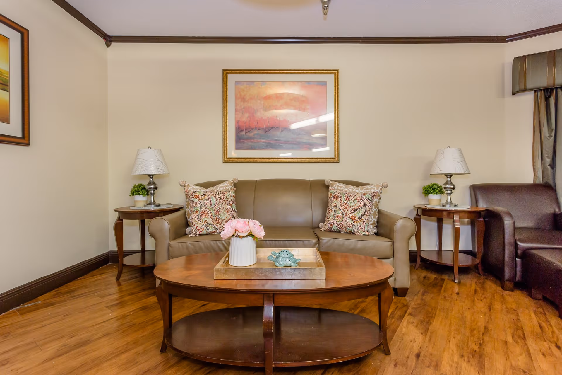 A cozy living room with a brown leather sofa adorned with two patterned cushions. On either side of the sofa are wooden side tables with white lamps and small green plants. A wooden coffee table in front of the sofa holds a tray with a white vase of pink flowers and decorative items. The room has wooden flooring, beige walls, and framed artwork hanging above the sofa.