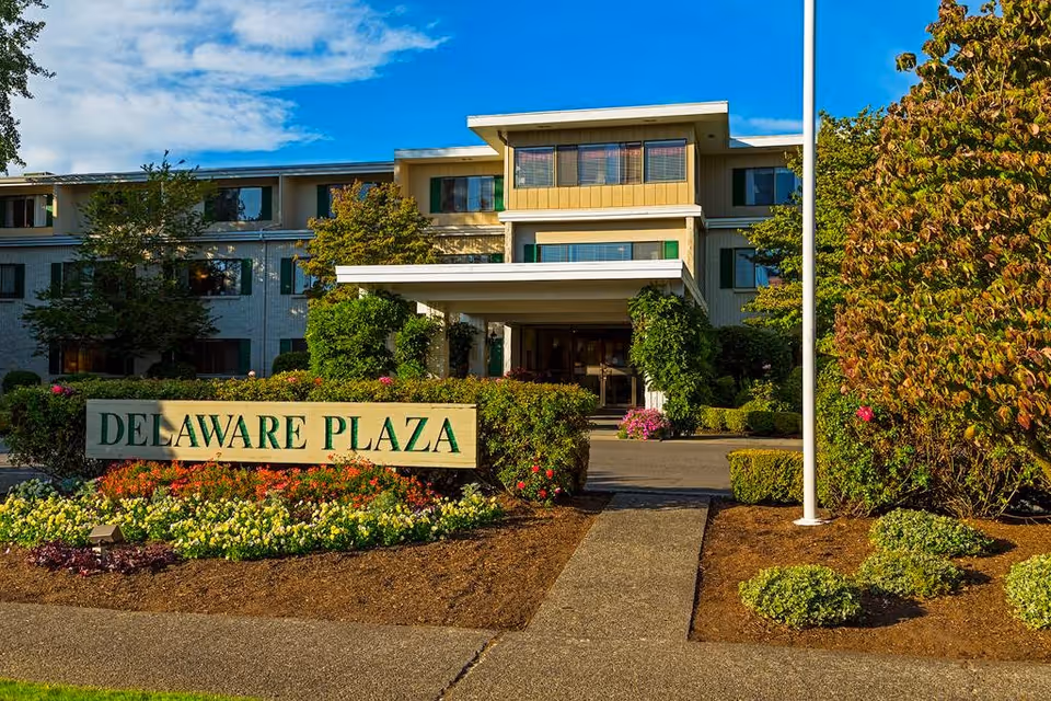Entrance of the Delaware Plaza assisted living building with a sign, landscaped flowerbeds, and a covered driveway.