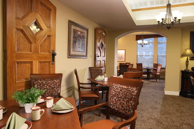 Interior view of a dining area in an assisted living facility with wooden tables and chairs, each table set with plates, mugs, and folded green napkins. The room features warm beige walls, decorative wooden doors with diamond-shaped windows, framed artwork, carpeted floors, and ceiling light fixtures including a chandelier.