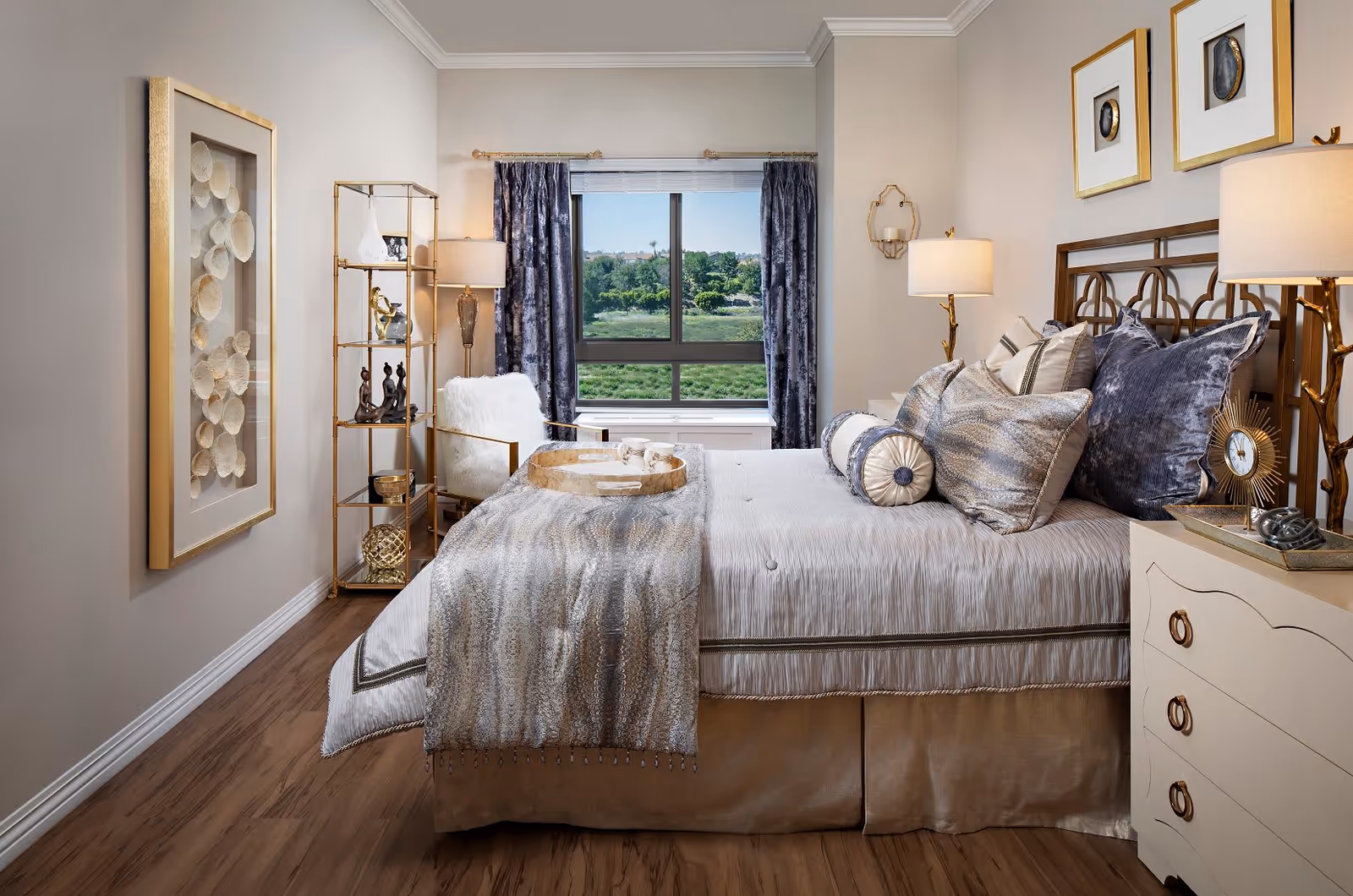 A well-decorated bedroom with a large bed adorned with multiple pillows and a textured throw blanket. There is a window with dark curtains showing greenery outside. The room features a white armchair, a gold and glass shelving unit with decorative items, two bedside tables with lamps, and framed artwork on the walls. The floor is wooden.